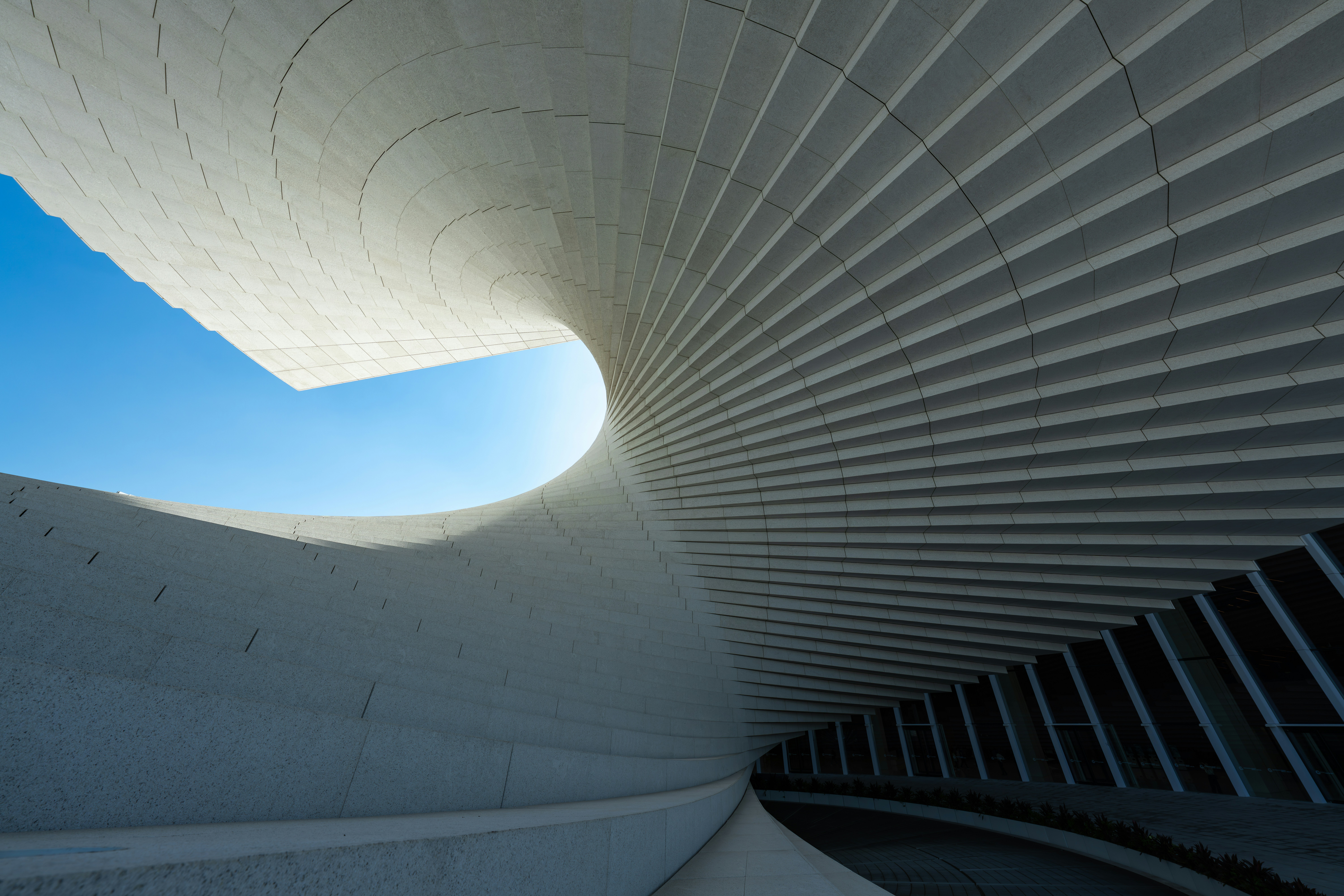 A low-angle shot of a modern building's interior, featuring a spiraling structure made of light-colored, textured concrete panels. A large, curved opening reveals a bright blue sky, contrasting with the shadowed, angled glass windows below.