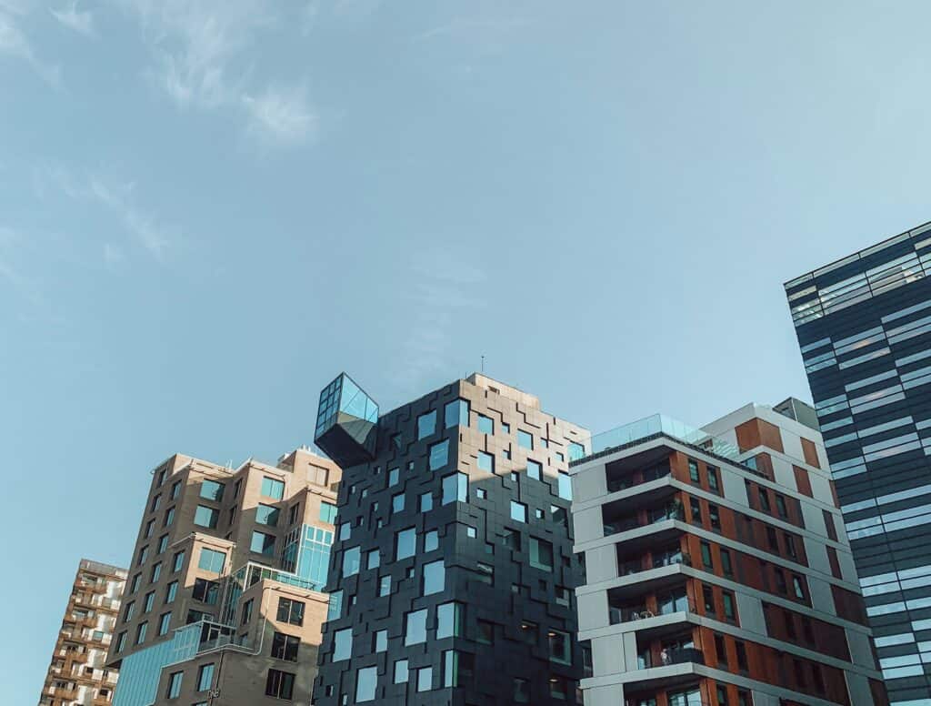 An upward view of several modern buildings with varied architectural styles, including a dark building with a protruding glass cube, against a clear blue sky.