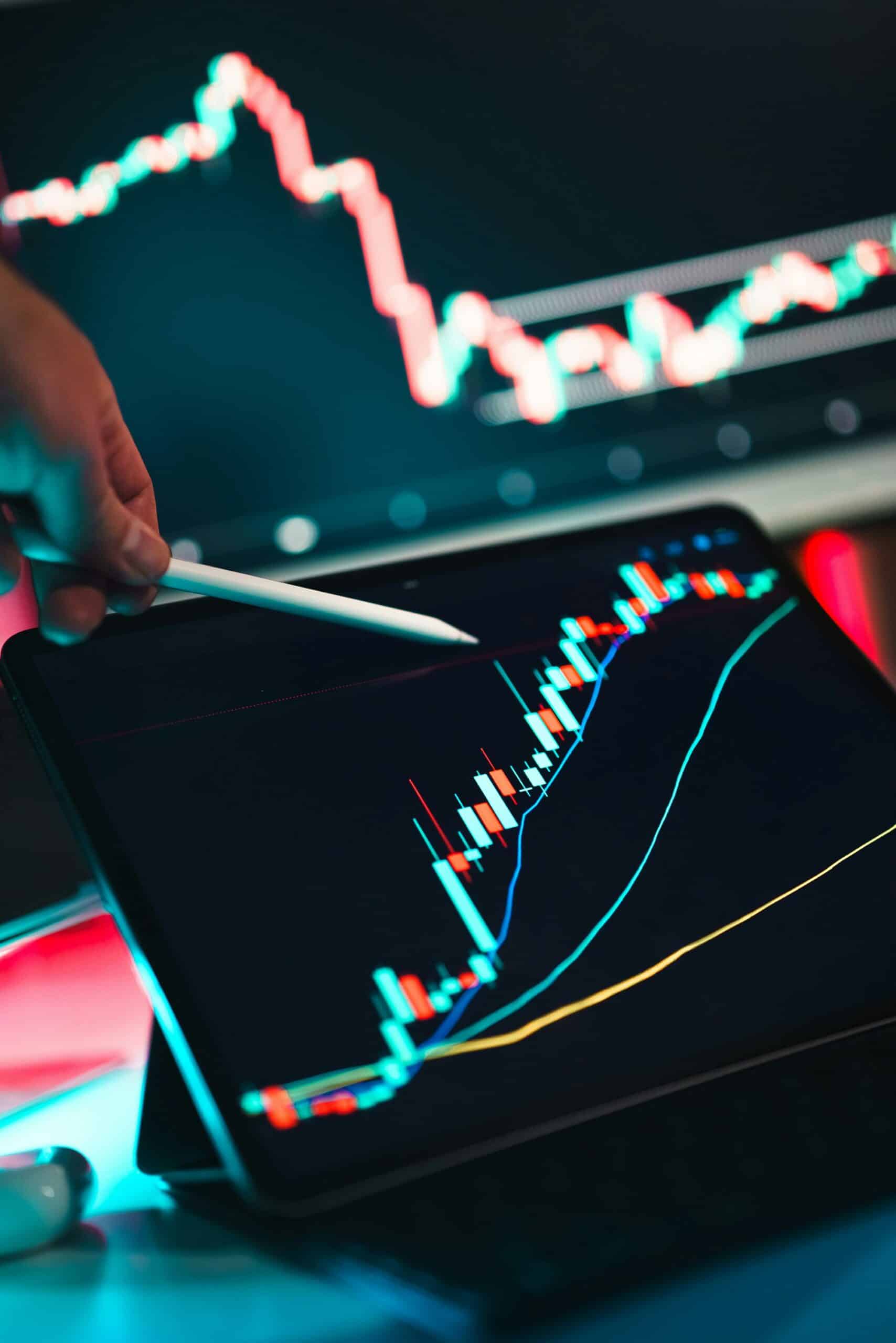 A hand holding a white stylus points at a tablet displaying a financial candlestick chart with red and teal bars. A blurred monitor in the background also shows a chart.
