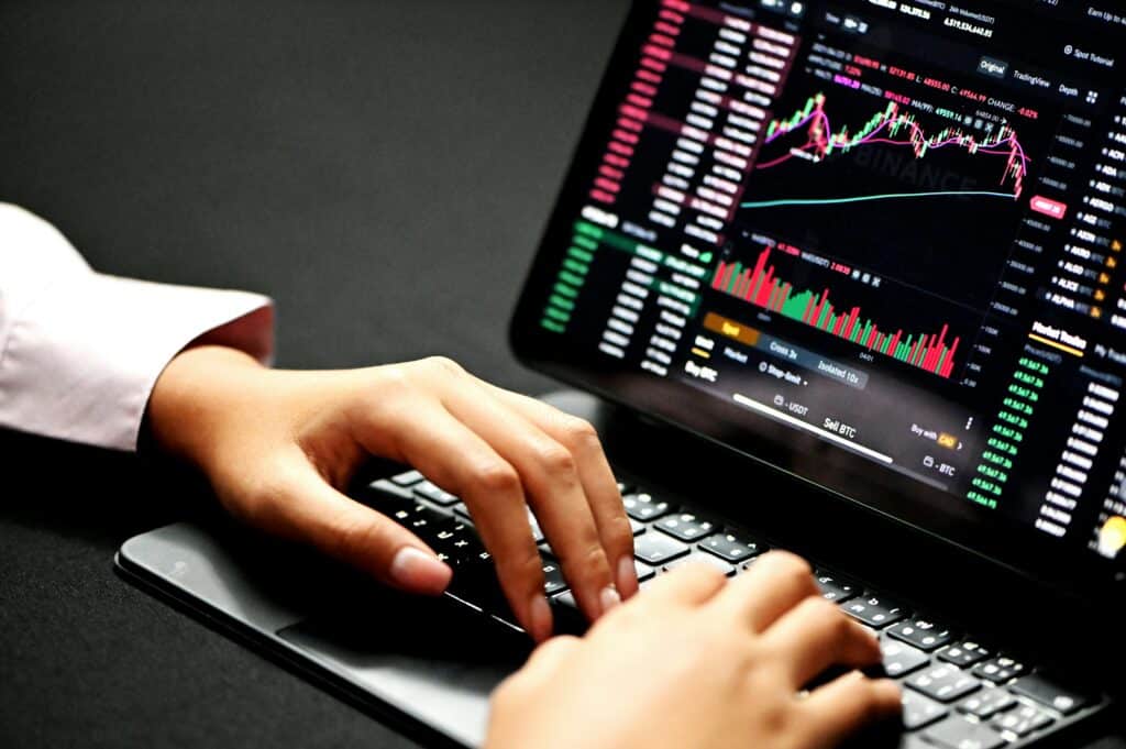 A person's hands typing on a laptop keyboard, with the screen displaying a cryptocurrency trading platform showing charts, market data, and buy/sell options for Bitcoin.