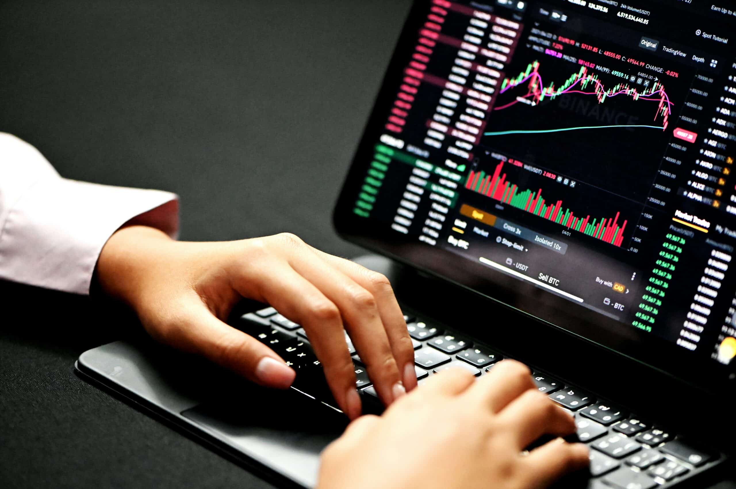 A person's hands typing on a laptop keyboard, with the screen displaying a cryptocurrency trading platform showing charts, market data, and buy/sell options for Bitcoin.